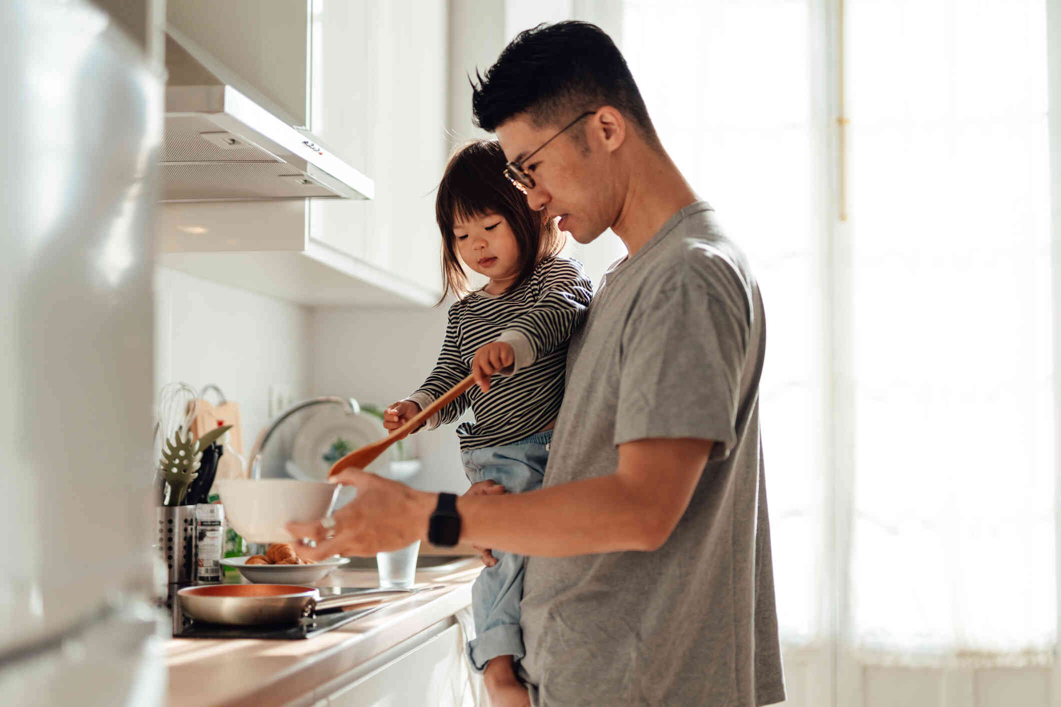 A dad stands in the kitchen while holding his young daughter on his hip as she holds a big wooden spoon and helps him put food in a bowl in his other hand.